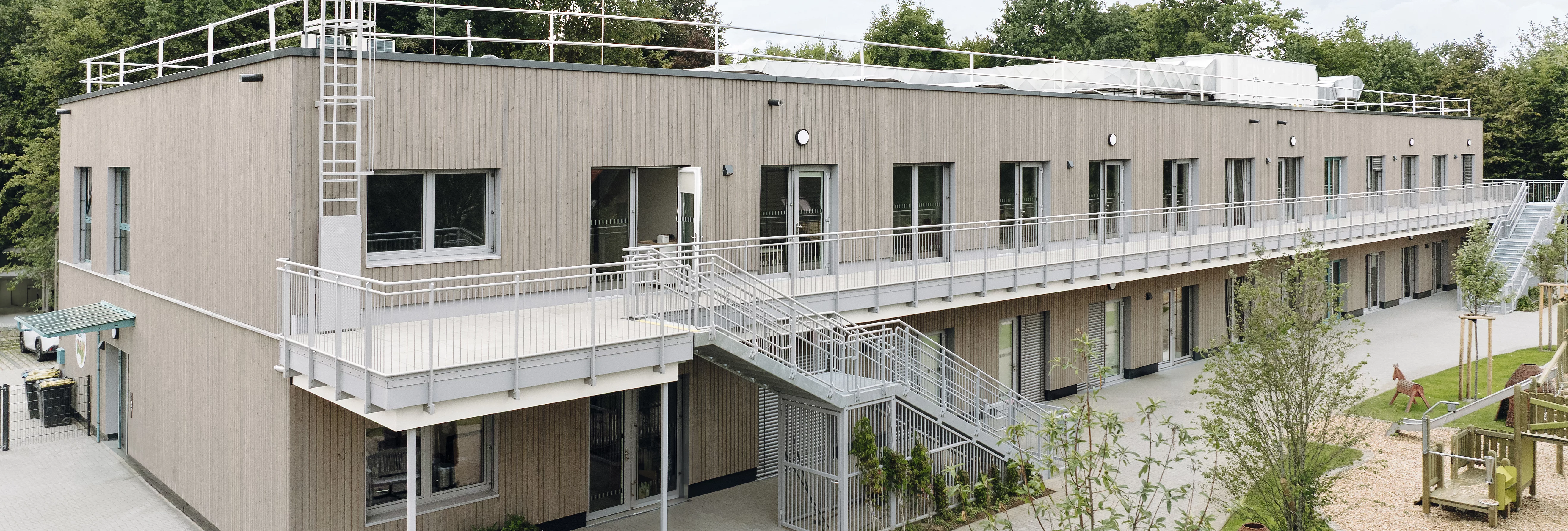 Zweigeschossiges Holzmodulgebäude mit Außentreppe, Laubengang, Spielplatz und begrüntem Außenbereich