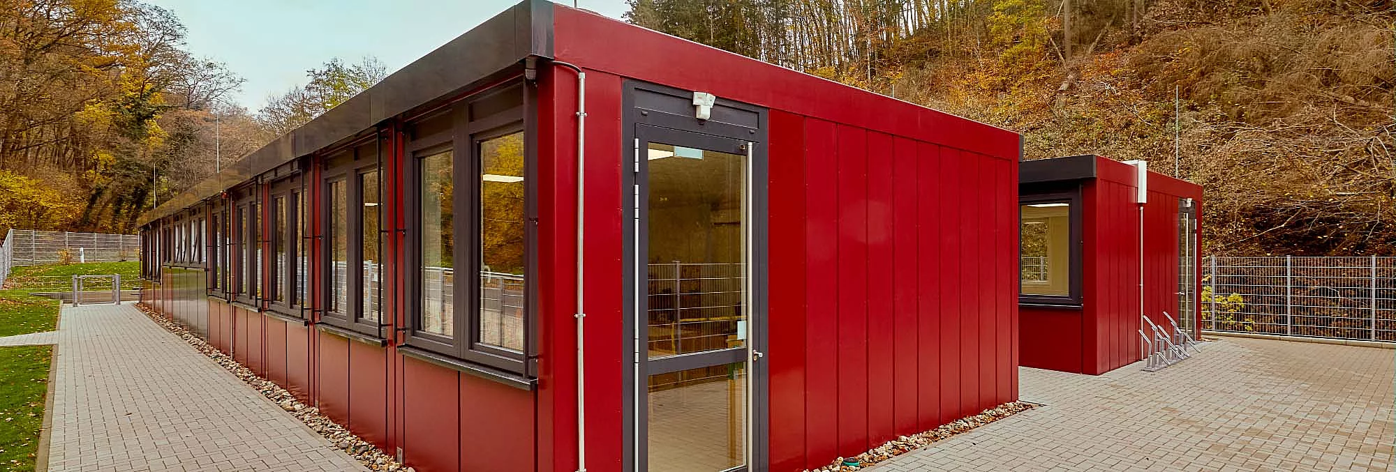 Rotes eingeschossiges Modulgebäude mit Glastür, Fensterreihen und gepflastertem Außenbereich