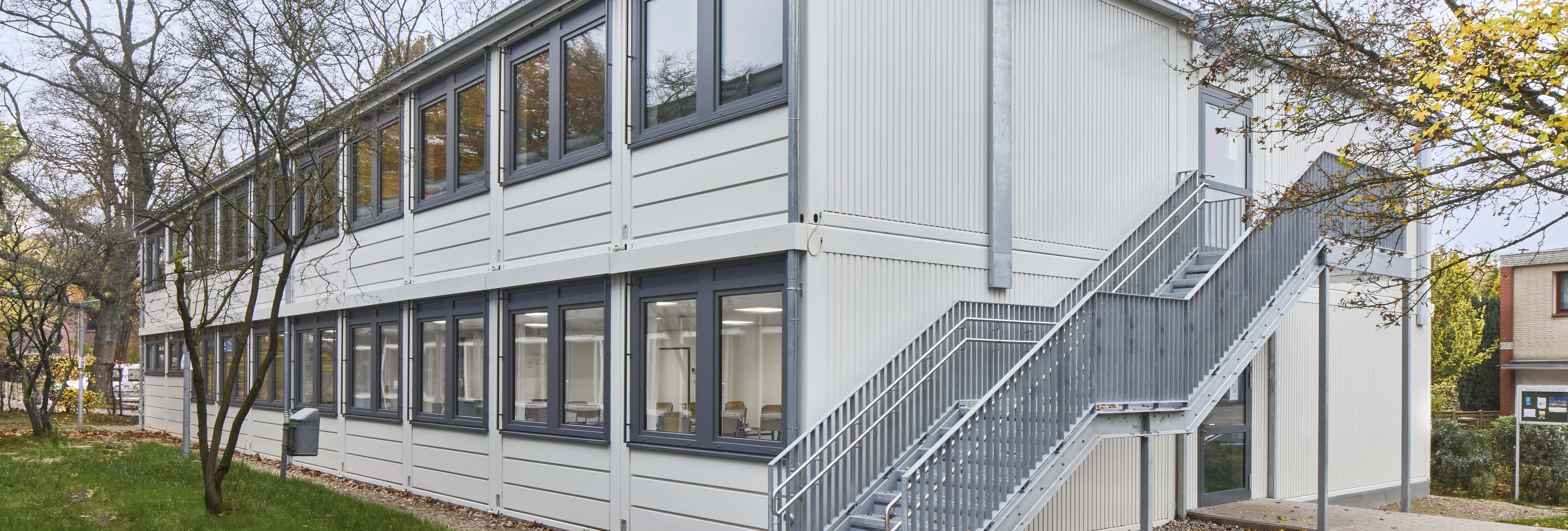 Zweigeschossige helle Containeranlage mit Außentreppe, Fensterreihen und Bäumen im Außenbereich