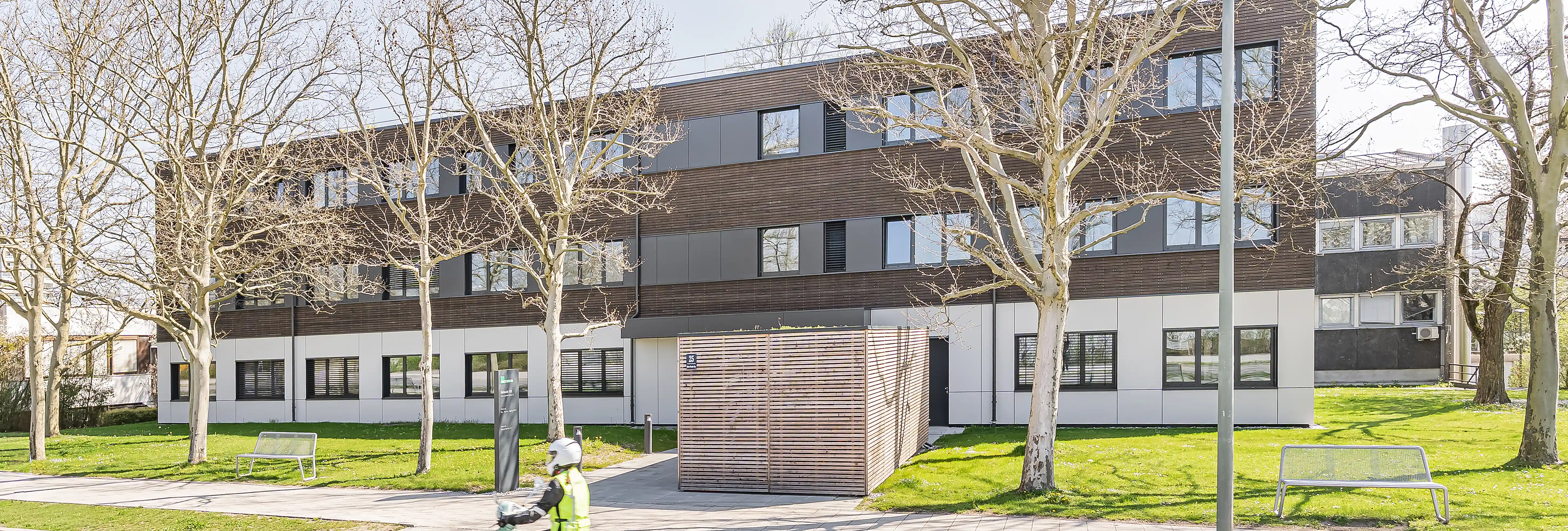 Dreigeschossiges Modulgebäude mit Holzlamellen-Fassade und hellem Sockel, eingebettet in parkähnliche Umgebung mit kahlen Bäumen und Rasenflächen