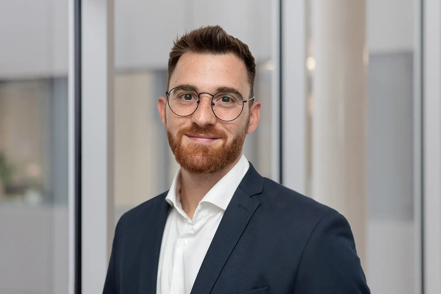 Porträt von Wasili Doulios mit runder Brille und dunkelblauem Sakko vor einer Glasfront