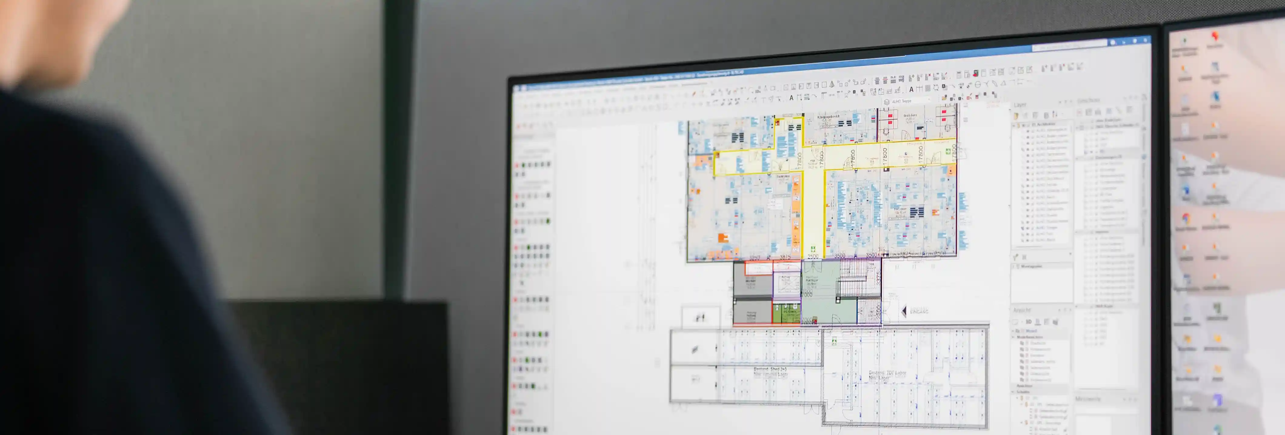Arbeitsplatz mit großem Monitor, auf dem ein technischer Gebäude- oder Hallenplan in einer CAD-Software dargestellt wird. Eine Person befindet sich unscharf im Vordergrund vor dem Bildschirm.