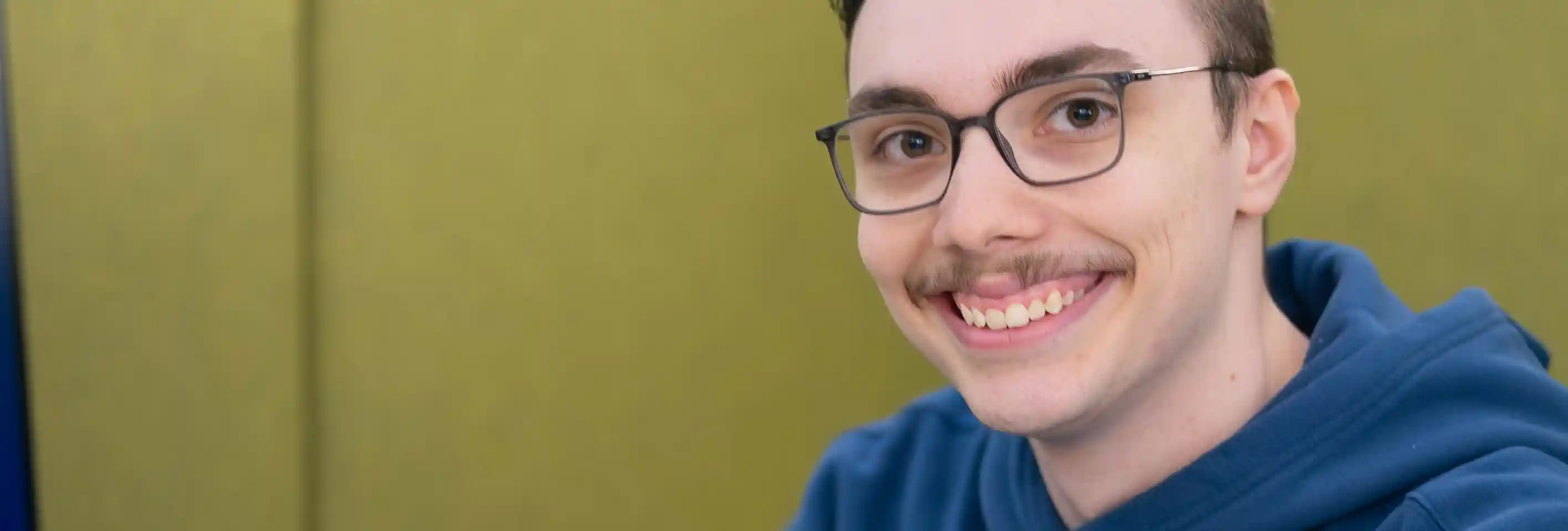 Nahaufnahme einer Person mit schwarzer Brille und blauem Kapuzenpullover. Die Person blickt in die Kamera. Der Hintergrund ist unscharf und in Grüntönen gehalten.