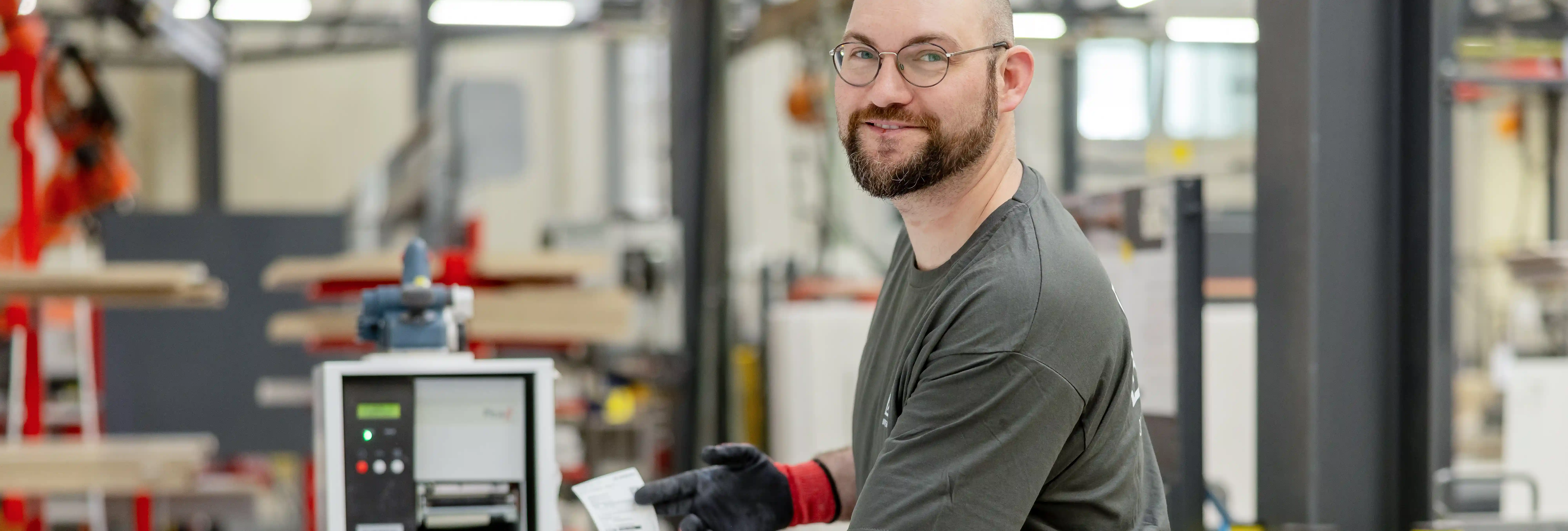 Person an einer Produktions- oder Verpackungsanlage in einer Industriehalle. Die Person trägt Arbeitshandschuhe und hält ein Etikett oder Dokument in der Hand. Im Hintergrund sind Maschinen, Fördertechnik und weitere Anlagenteile einer Fertigungsumgebung zu sehen.