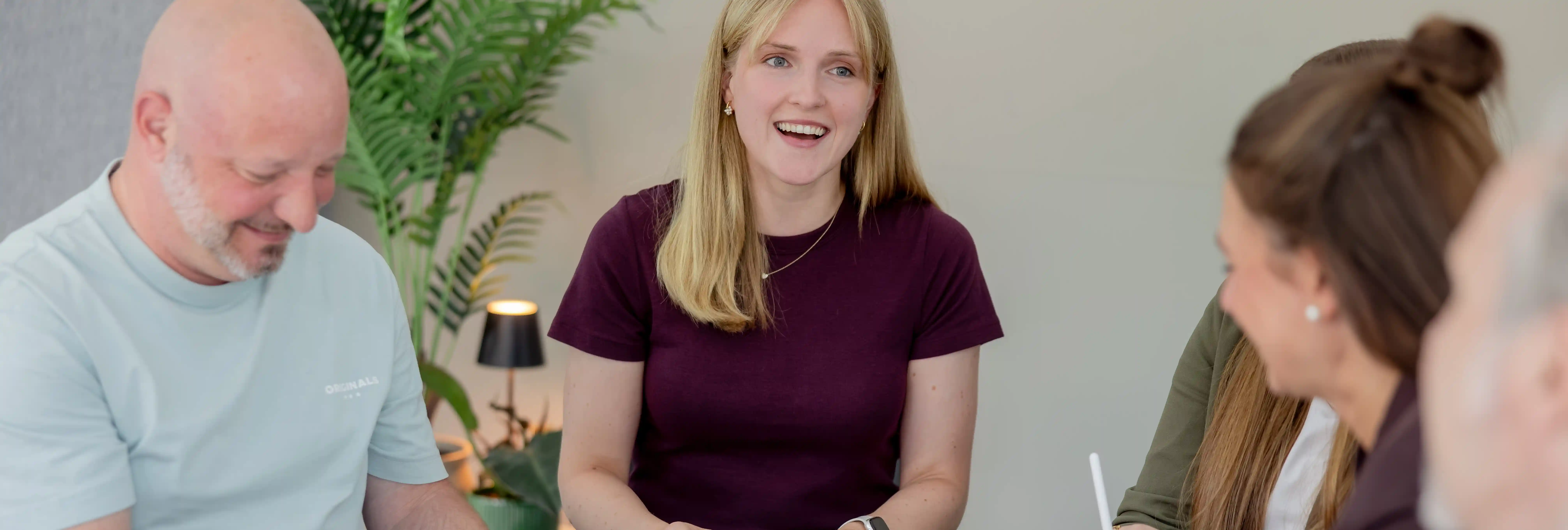 In einem hellen Besprechungsraum sitzen mehrere Personen an einem Tisch. Eine Person in der Bildmitte spricht oder präsentiert, während die anderen Personen der Runde zugewandt sind. Im Hintergrund sind Zimmerpflanzen und eine kleine Tischleuchte zu sehen, die dem Raum eine moderne Arbeitsatmosphäre verleihen. Die Aufnahme zeigt eine kollaborative Besprechungssituation in einem professionellen Büroumfeld.