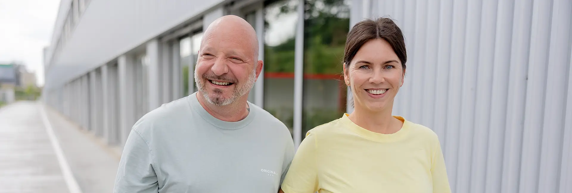 Zwei Personen stehen vor einer modernen Gebäudefassade mit großen Fensterflächen und blicken in die Kamera. Die Aufnahme entstand im Außenbereich bei Tageslicht.