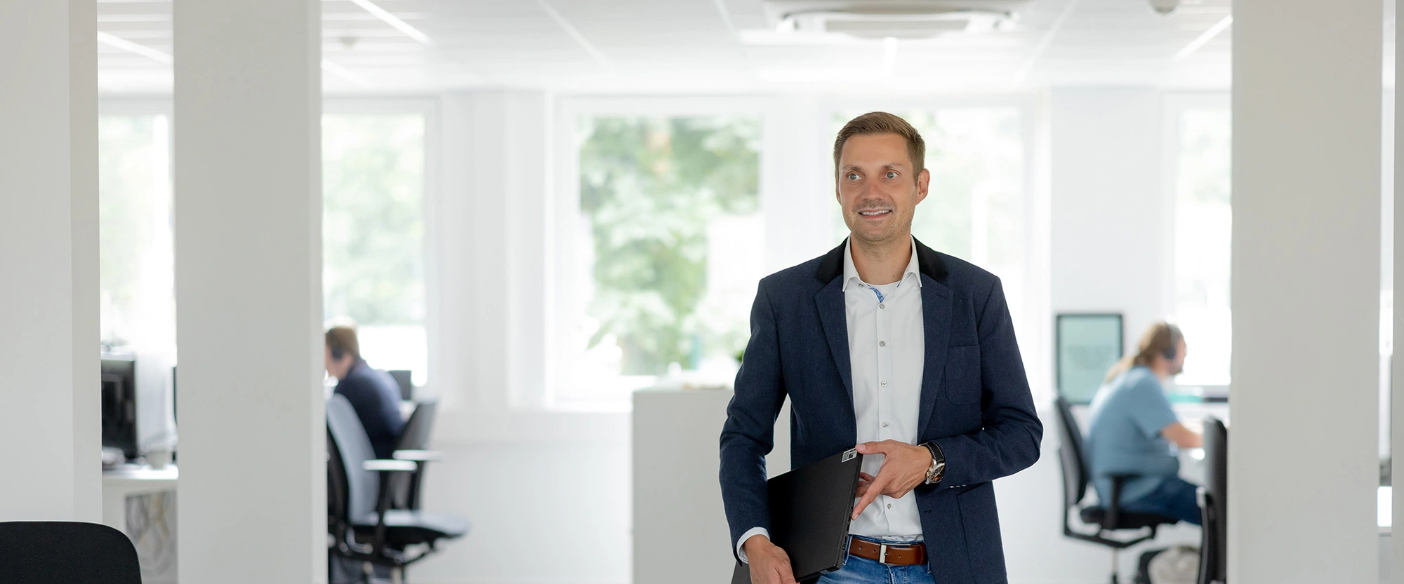 Mann im Sakko geht mit Laptopmappe durch ein helles Büro mit Arbeitsplätzen im Hintergrund