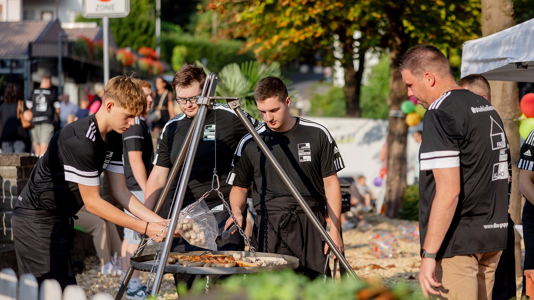Vier junge Männer in schwarzen Trikots grillen Fleisch unter freiem Himmel bei Sonnenlicht. Im Hintergrund sieht man Menschen und Bäume.