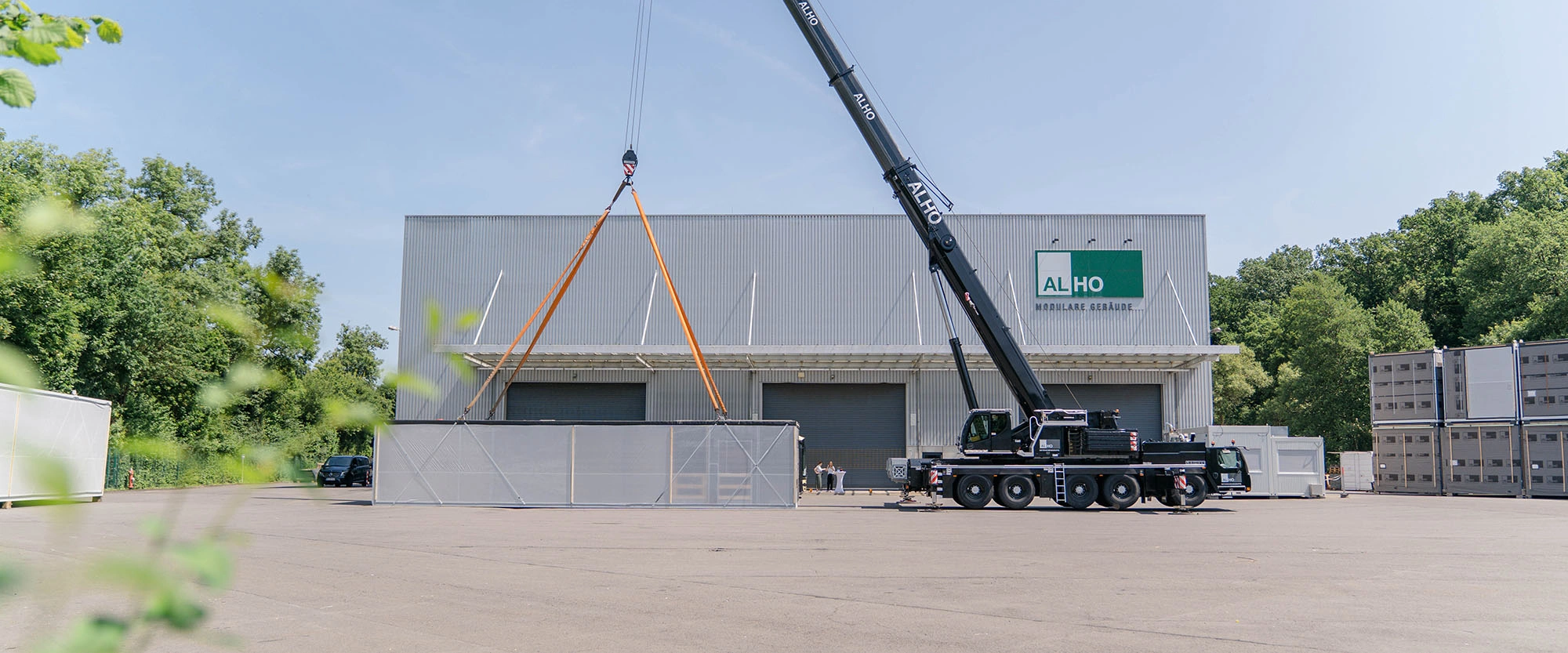 Montage eines modularen Raummoduls mit Kran auf Werksgelände, im Hintergrund Industriehalle mit Schriftzug „ALHO Modulare Gebäude“ sowie gestapelte Module rechts.