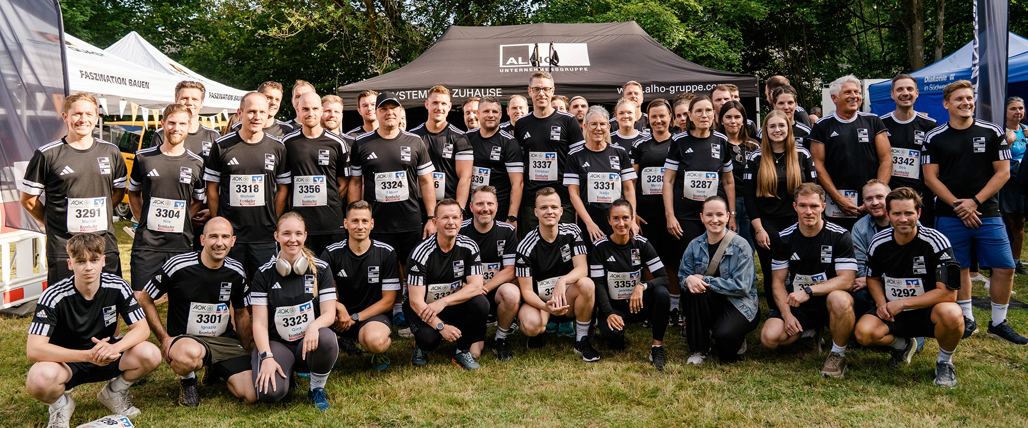 Eine Gruppe von Menschen in schwarzen Sporttrikots posiert fröhlich nach einem Laufwettbewerb vor einem Zelt im Freien. Sie tragen Startnummern und stehen oder knien auf Gras.