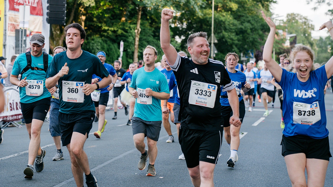Menschen laufen bei einem Stadtlauf, jubelnde Teilnehmerinnen und Teilnehmer mit Startnummern und WDR-Shirts im Vordergrund.