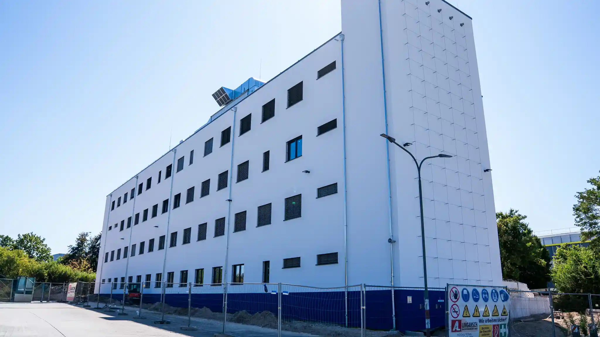 Außenansicht des Erweiterungsbaus der IGS Europa in Mainz-Hechtsheim. Das viergeschossige Schulgebäude mit heller Fassade und zahlreichen Fenstern erstreckt sich entlang einer Straße. Vor dem Gebäude befinden sich Bauzäune sowie Hinweisschilder zur Baustellensicherheit. Die Aufnahme entstand bei sonnigem Wetter, umgeben von Bäumen und Grünflächen. Das Gebäude wurde in modularer Bauweise realisiert und kurz vor der Inbetriebnahme fotografiert.