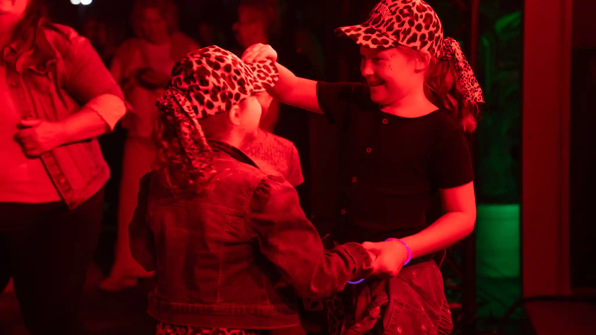 Zwei Kinder mit gemusterten Kappen halten sich an den Händen und tanzen auf einer Veranstaltung mit roter Bühnenbeleuchtung. Weitere Gäste befinden sich im Hintergrund.