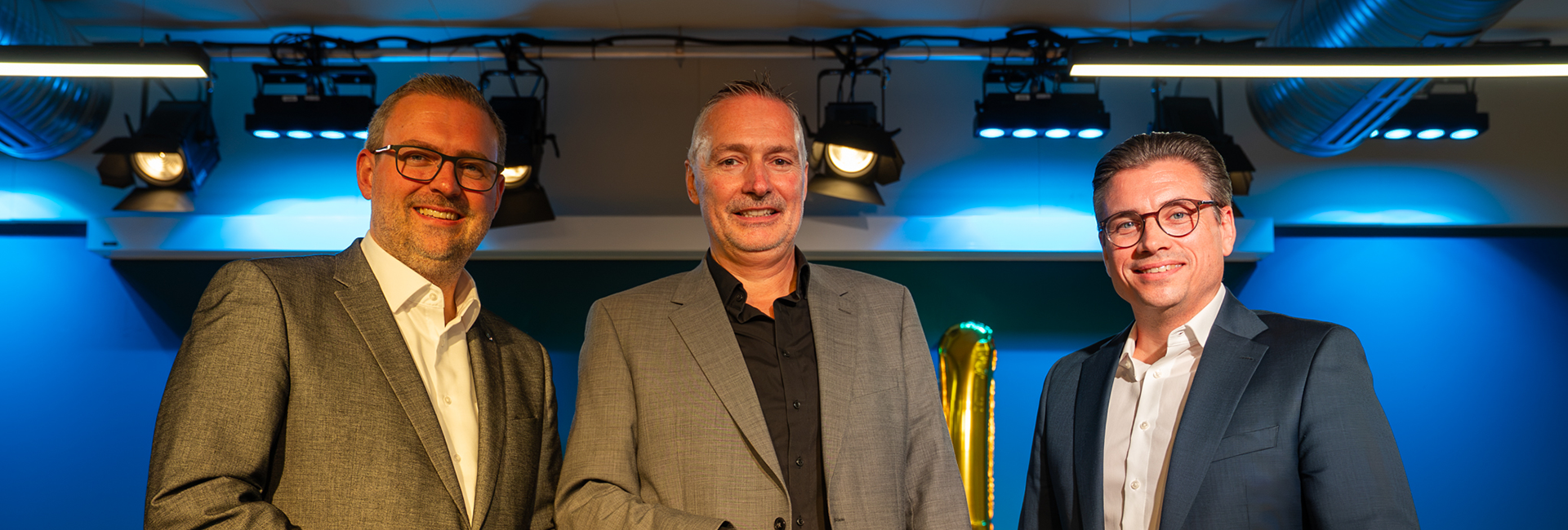 Bürgermeister Erik Lierenfeld, Schulleiter Jörg Hustede und Sebastian Trautermann stehen auf einer Bühne mit blauer Beleuchtung und blicken in die Kamera. Im Hintergrund sind Bühnenstrahler und dekorative Ballons zu sehen, aufgenommen im Rahmen der Eröffnungsfeier der Realschule Dormagen. 