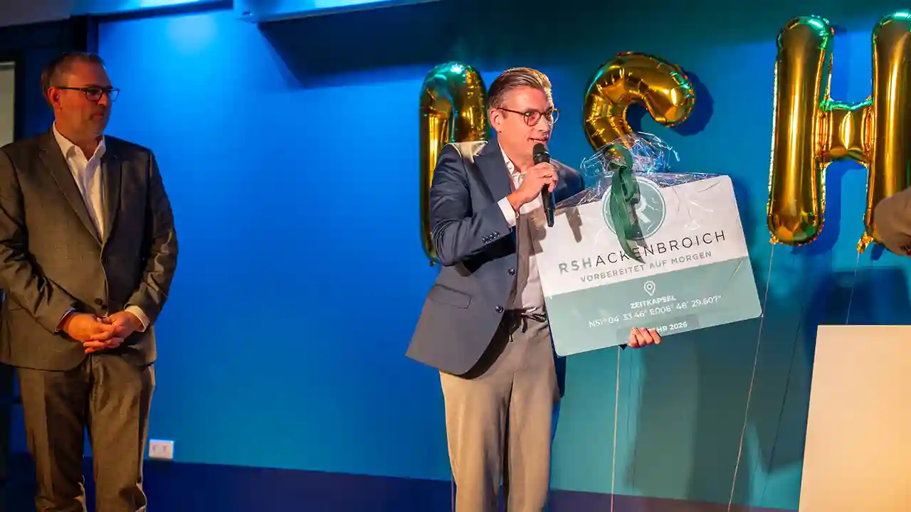 Person hält während einer Feier ein Türschild mit den Koordinaten einer Zeitkapsel und spricht in ein Mikrofon. Im Hintergrund sind Luftballons und das Logo von RSHACKENBROICH zu sehen.