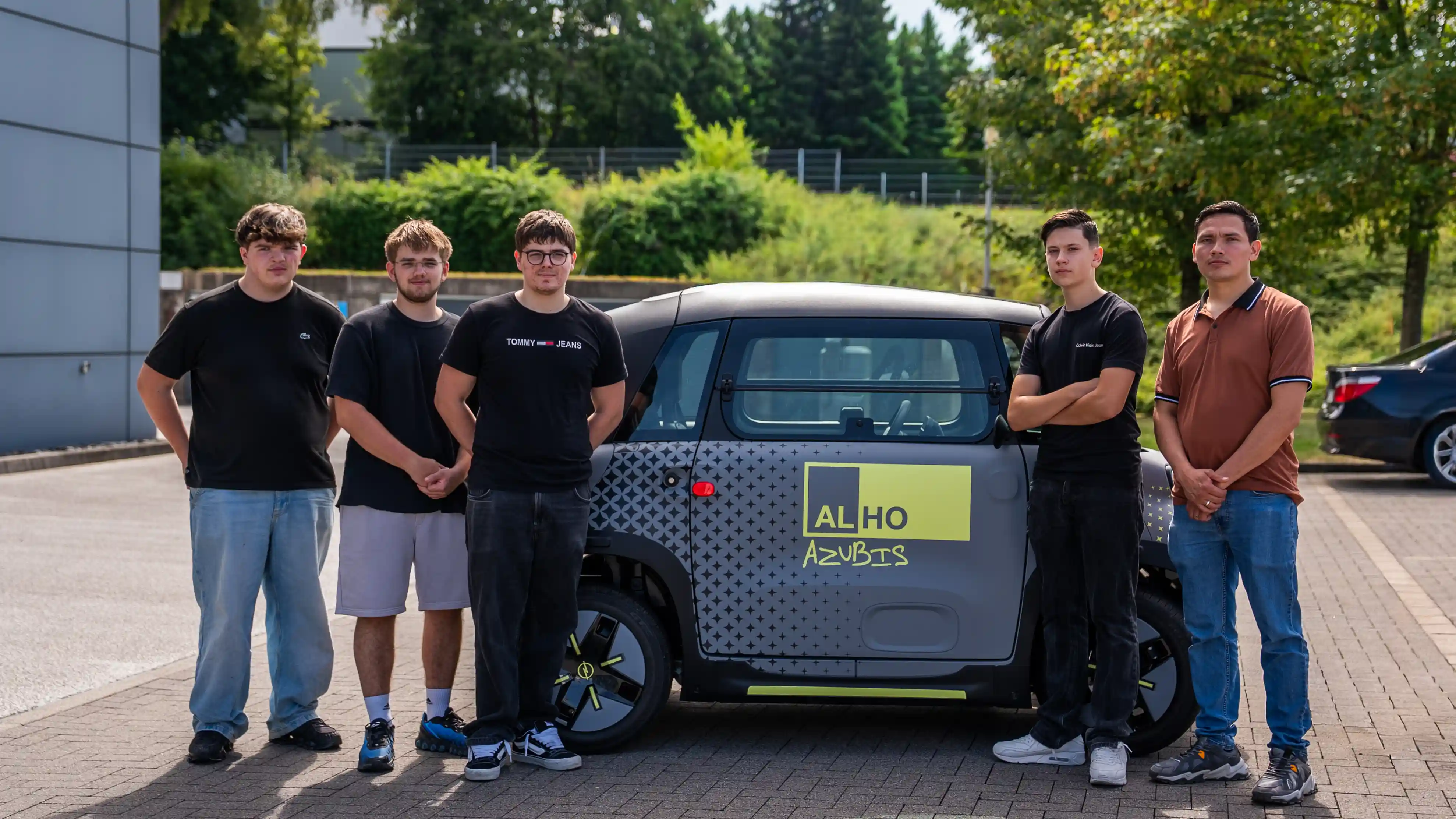 Fünf ALHO Auszubildende stehen vor einem Kleinwagen mit ALHO Azubis Beschriftung auf dem Firmenparkplatz