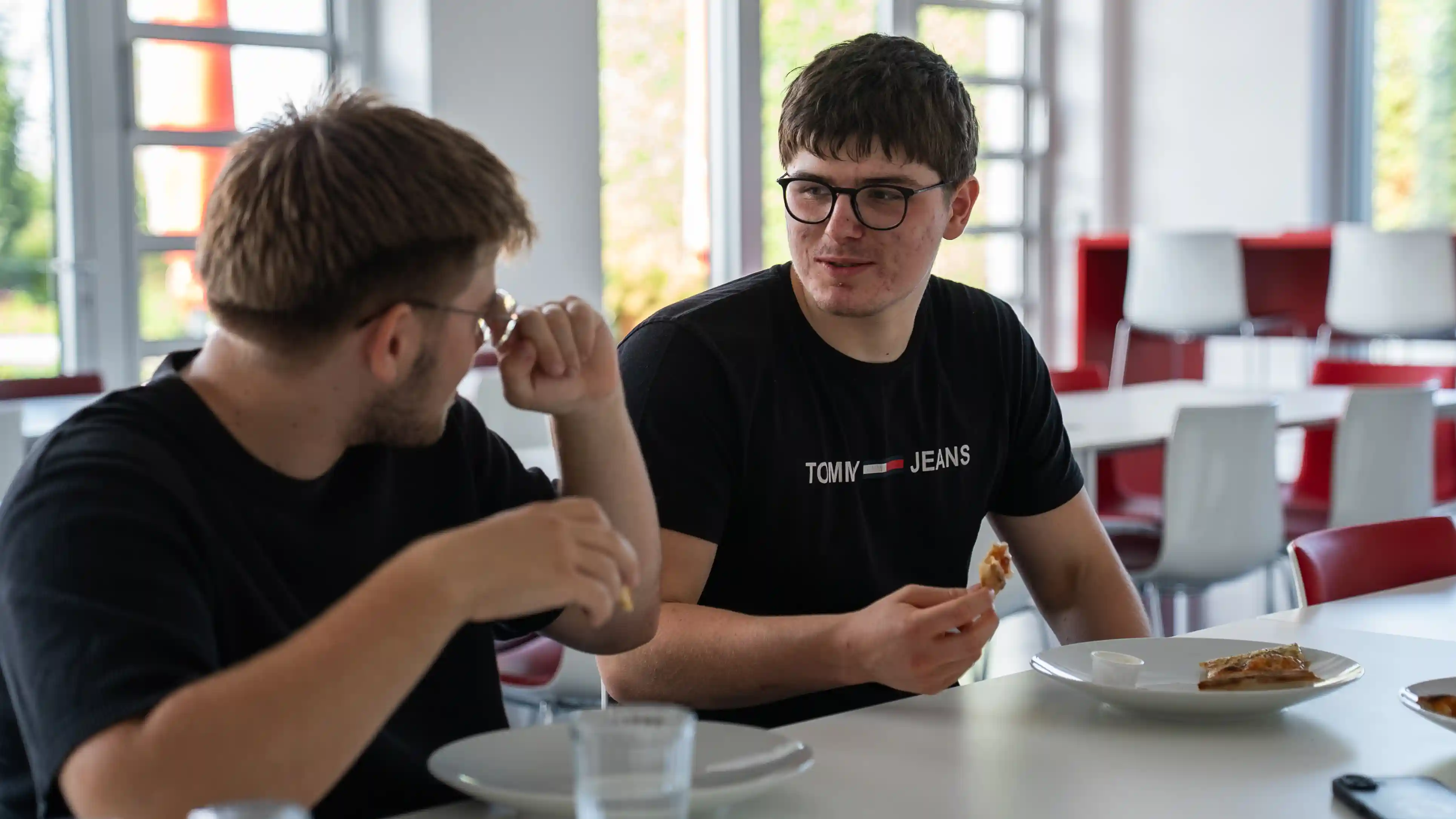 Zwei junge Männer sitzen im Pausenraum am Tisch und essen gemeinsam Pizza